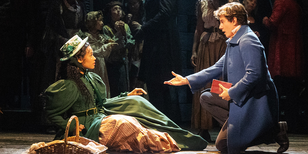 A woman in a green dress sits on stage, while a man in a blue coat kneels, offering a book.