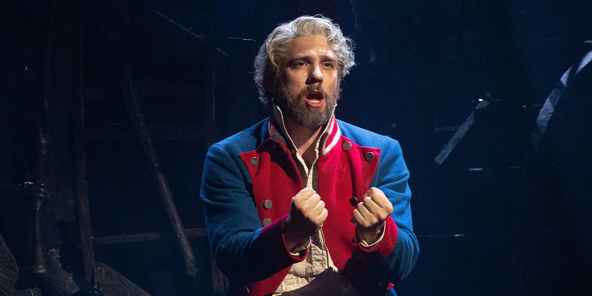 A man with grey hair and beard passionately sings, wearing a blue and red jacket, against a dark background.