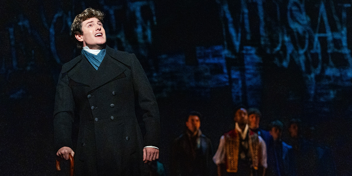 A young man in a dark coat sings passionately on stage, with a group of performers in the background.