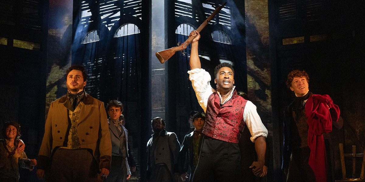 A man in a red waistcoat raises a rifle, surrounded by five other performers in period costumes.