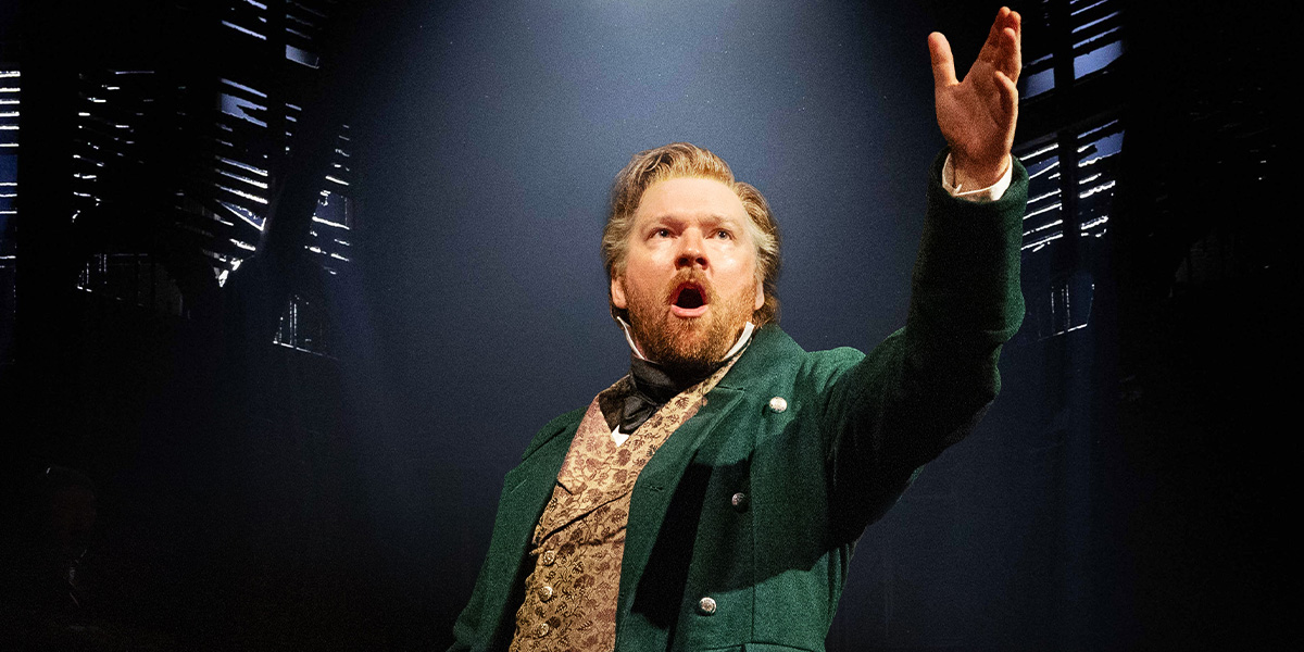 Killian Donnelly as Jean Valjean, wearing a dark coat, stands against a blurred theatre backdrop.