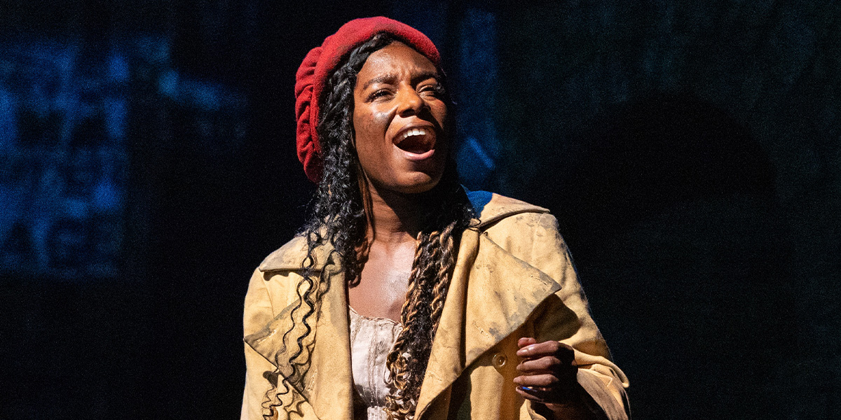 A woman with long, curly hair and a red beret sings passionately, wearing a tattered yellow coat.
