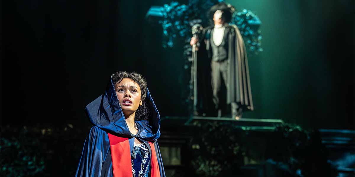 Beatrice Penny-Touré as Christine Daaé looks up, wearing a dark cloak, with a figure behind her.