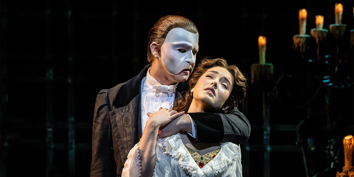 The Phantom, in a white mask and dark coat, embraces Christine, who wears a white gown, against a dark backdrop.