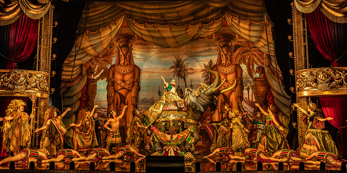 A vibrant theatrical scene featuring performers in elaborate costumes, an elephant, and dramatic backdrops.