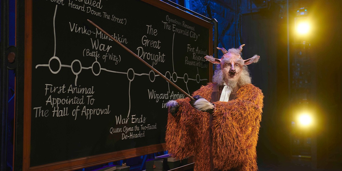 David McKechnie as Fiyero stands centre stage in a green costume, with dramatic lighting and a dark background.