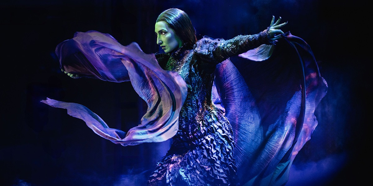 Emma Kingston as Elphaba, dressed in a green costume, stands confidently on stage with dramatic lighting.