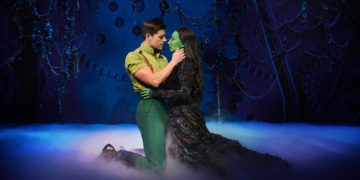 Jordan Litz as Fiyero and Emma Kingston as Elphaba pose together, wearing vibrant costumes against a dramatic backdrop.