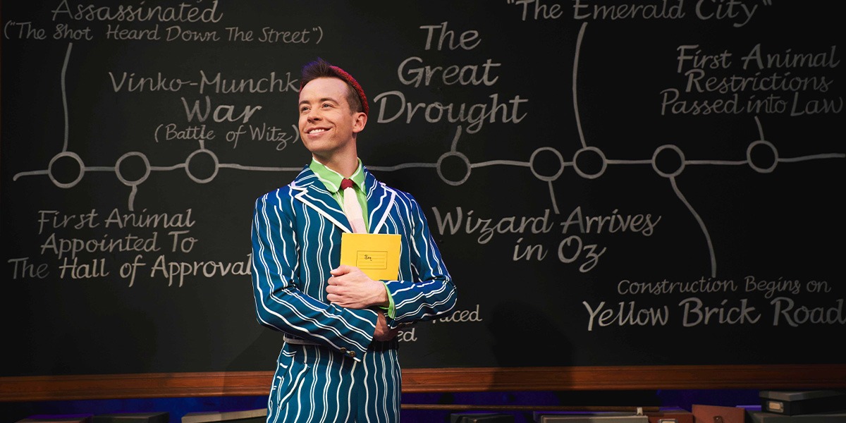 A performer in a green costume poses dramatically against a dark stage backdrop with soft lighting.