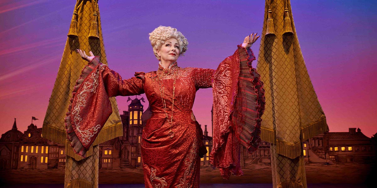 Sarah Ingram as Elphaba, wearing a green dress, stands on stage with dramatic lighting in the background.