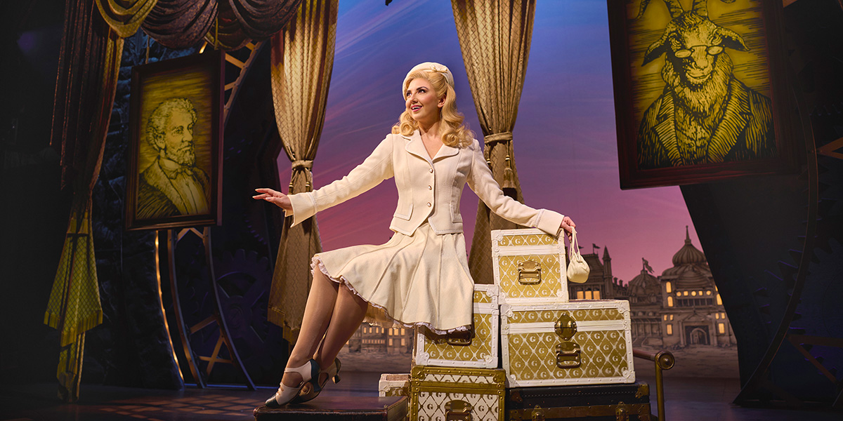 Zizi Strallen as Glinda, wearing a sparkling gown, smiles brightly against a theatrical backdrop.
