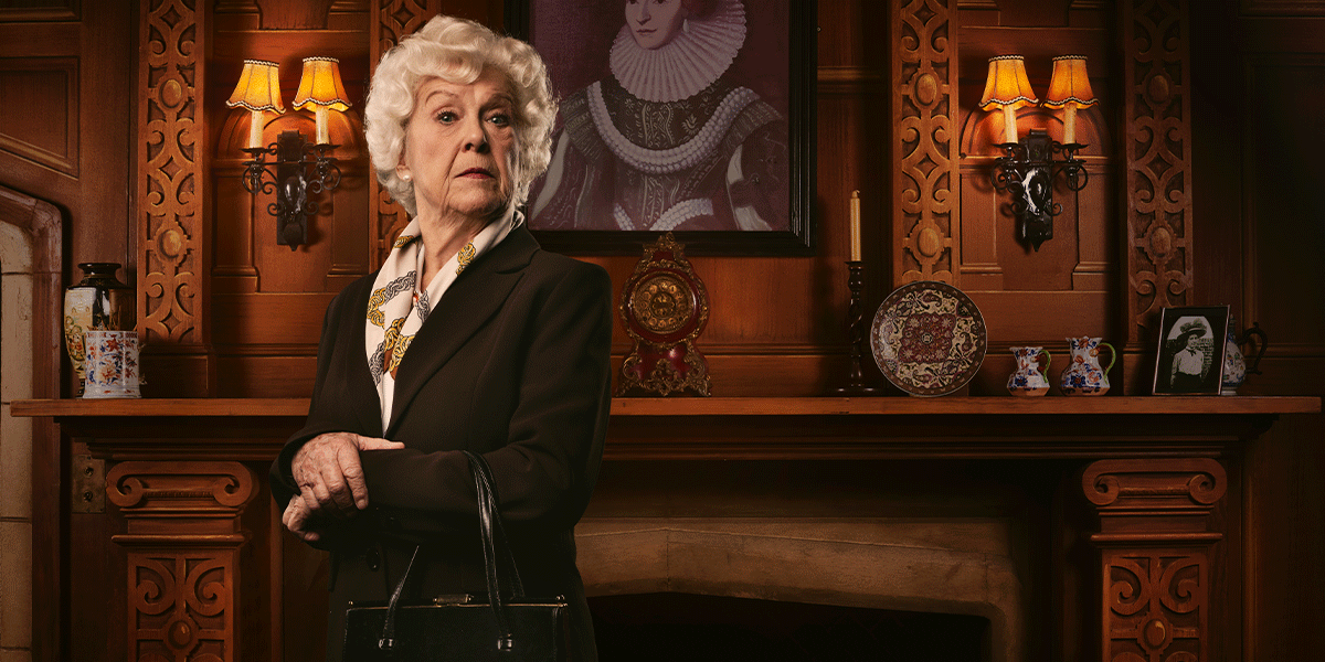 An elderly woman with curly white hair stands confidently, wearing a dark suit and holding a handbag.