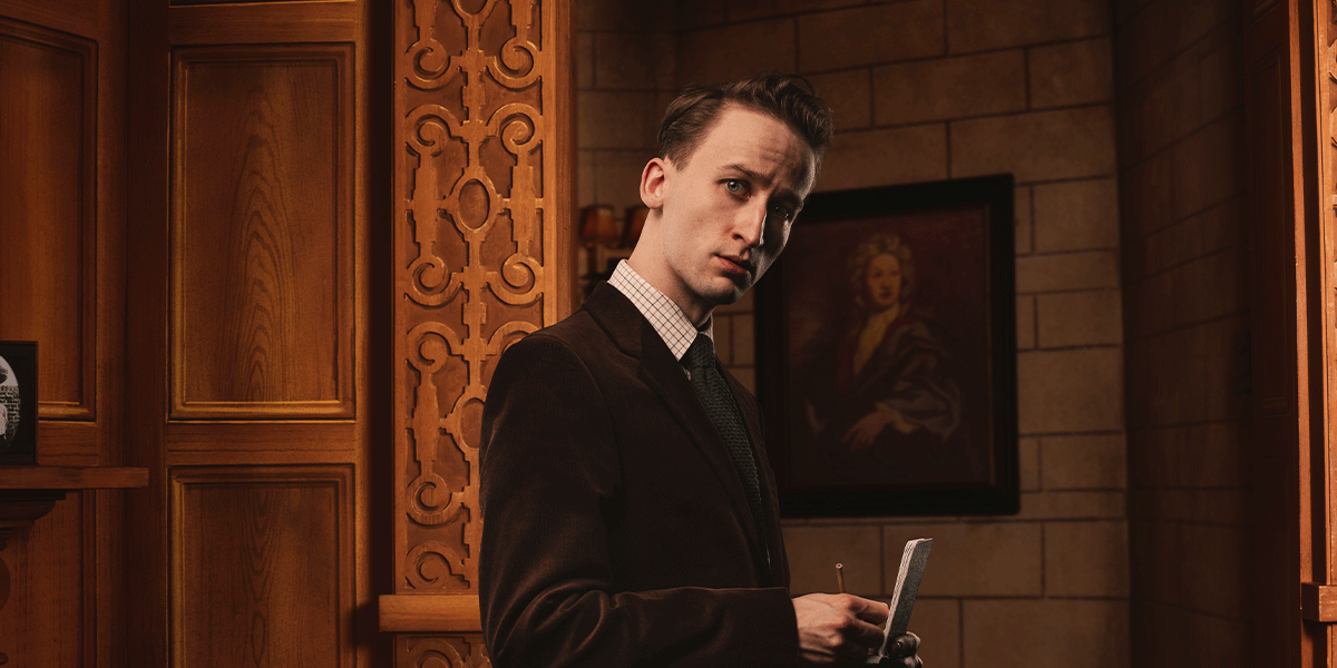 A man in a brown velvet suit holds a notepad, looking thoughtfully against a wooden backdrop.