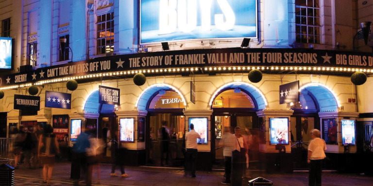 Piccadilly Theatre entrance illuminated with blue lights, featuring posters and people walking by at night.