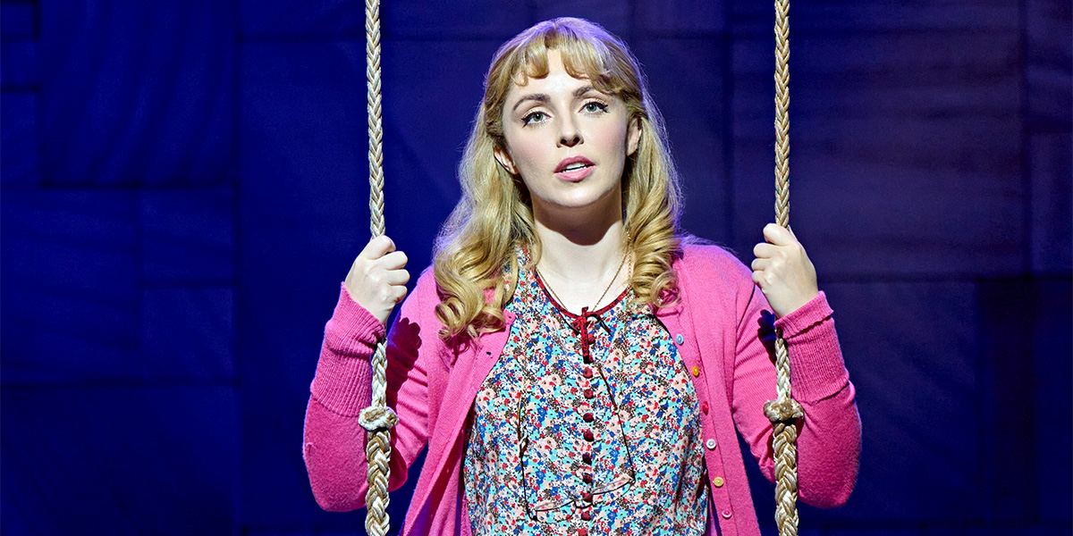 A woman with long, wavy hair wears a pink cardigan and floral blouse, holding a rope swing.