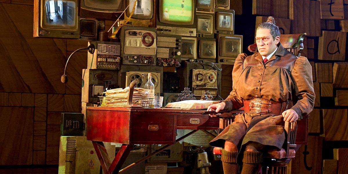 Jon Robyns as Miss Trunchbull, wearing a dark suit and stern expression, stands on stage in a theatrical setting.