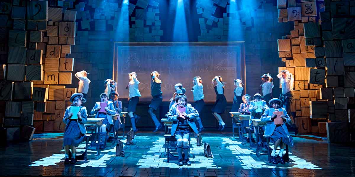 Children wearing grey school uniform sit at wooden desks with vibrant stage lighting and wooden blocks.