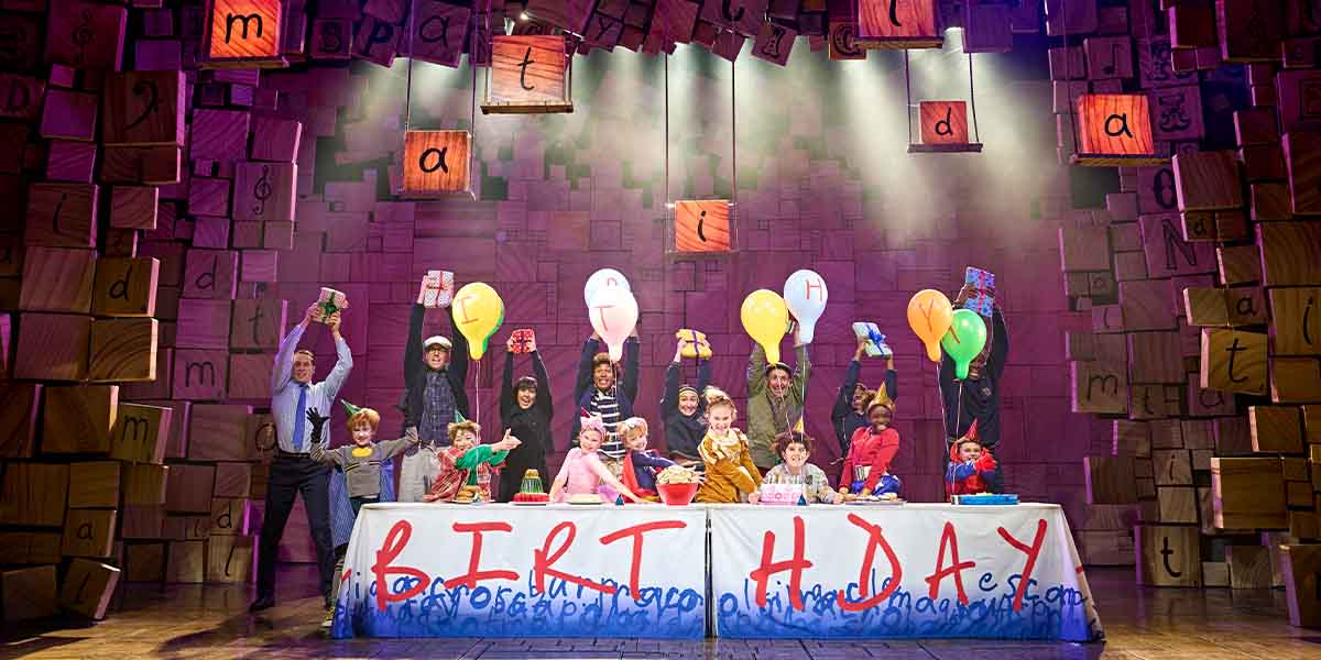 A group of children and adults celebrate a birthday on stage, holding balloons and gifts.