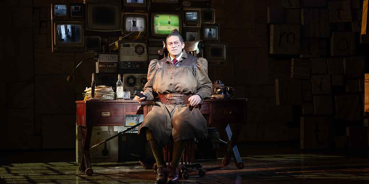 A person wearing a brown dress with a belt sits on a chair surrounded by vintage televisions and papers.