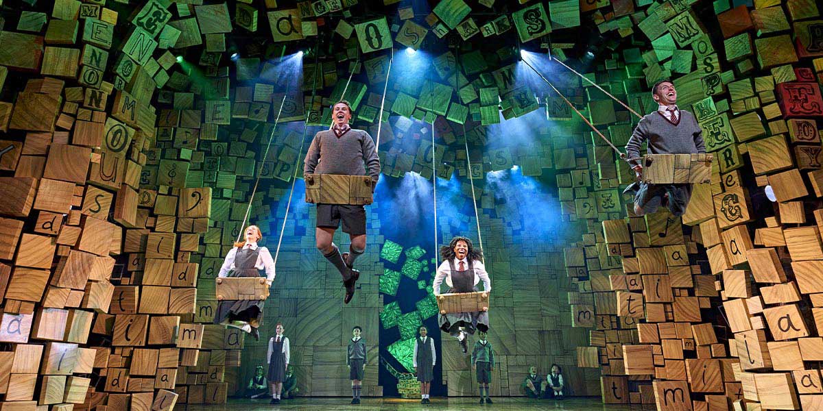 Edie McCaig as Matilda stands centre stage, surrounded by the cast in colourful costumes, smiling.
