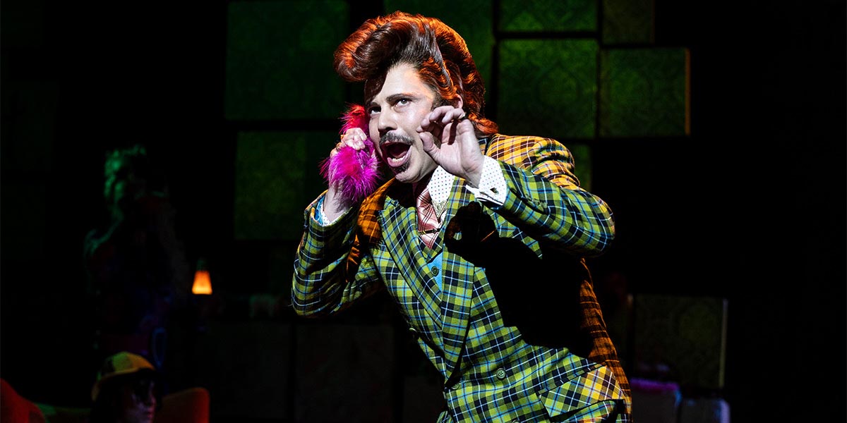 Neil McDermott as Mr Wormwood, wearing a bright suit, poses dramatically on stage in Matilda The Musical.