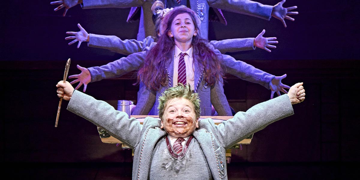 A scene from Matilda The Musical featuring actors in vibrant costumes performing on a brightly lit stage.