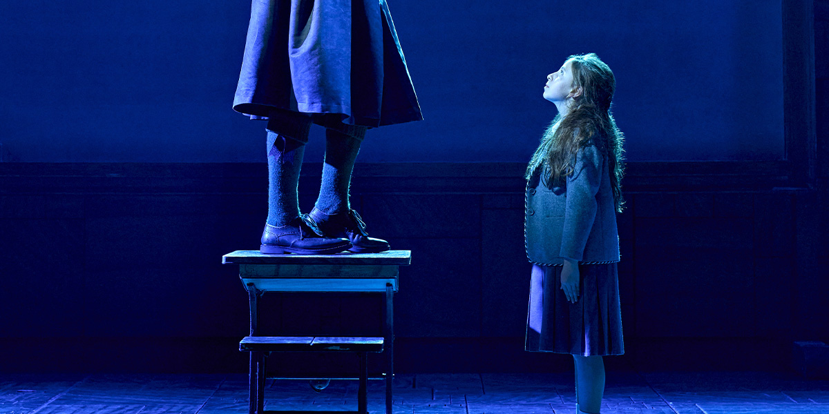 A girl looks up at a figure standing on a stool, illuminated by blue stage lighting.