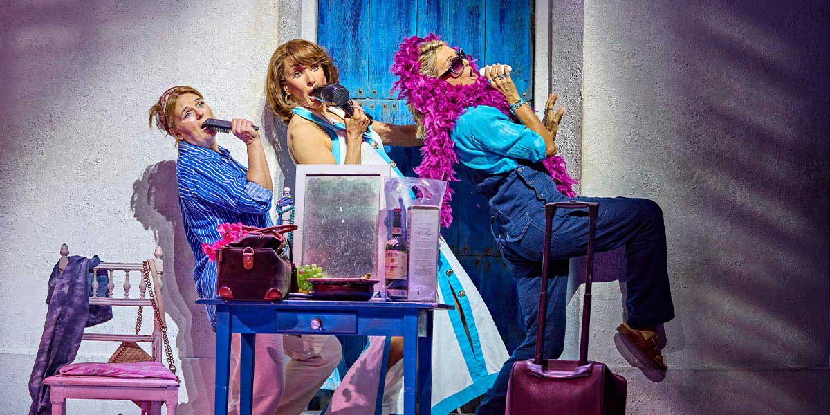 A scene from Mamma Mia! featuring performers in vibrant costumes, dancing joyfully on a stage with bright lighting.