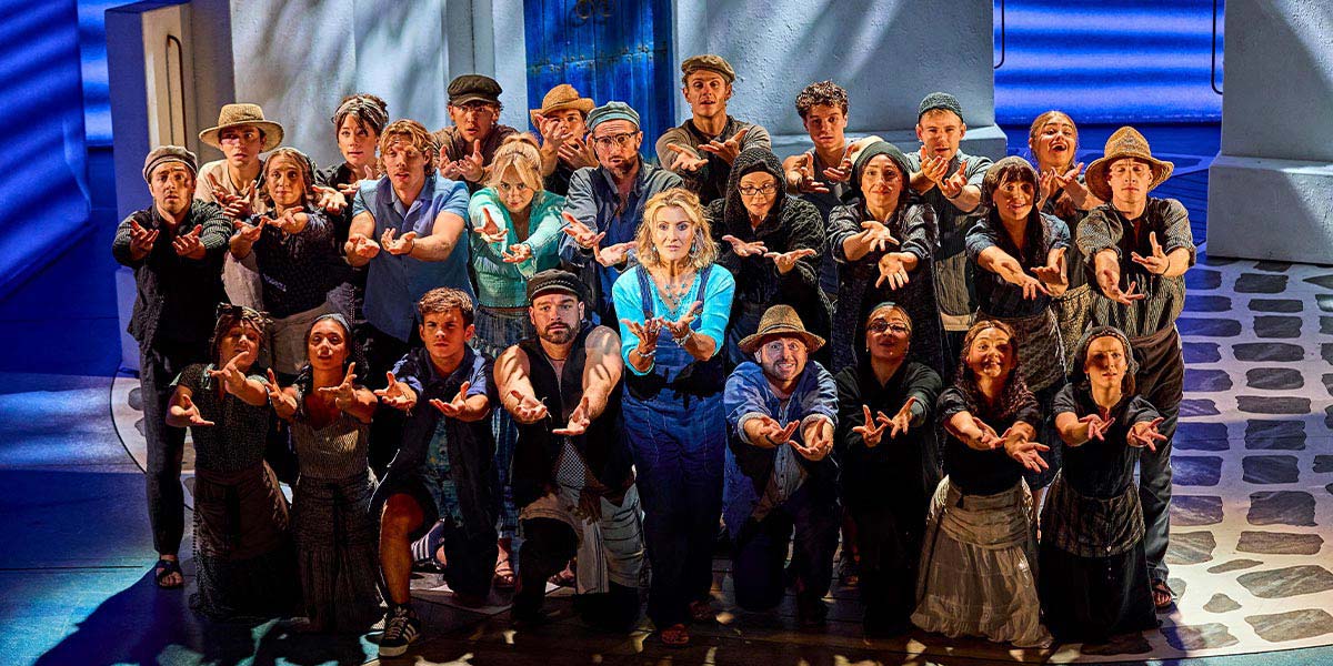 A vibrant scene from Mamma Mia! featuring actors in colourful costumes performing energetically on stage.