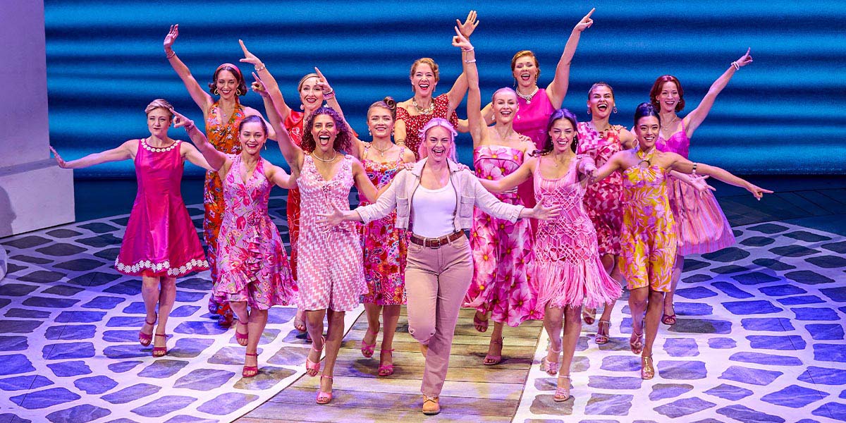The cast of Mamma Mia! performs on stage, featuring vibrant costumes and a lively backdrop.