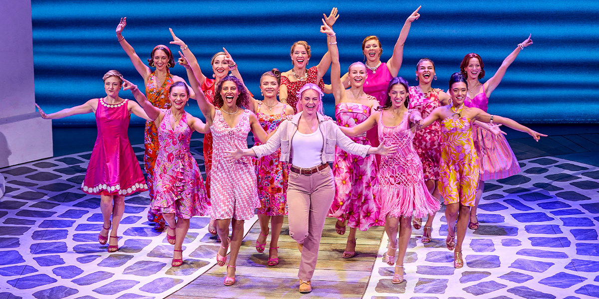 A group of women in vibrant pink dresses joyfully dance together on stage, smiling and posing.