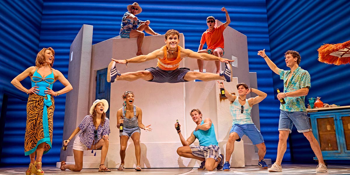 A scene from Mamma Mia! featuring performers in vibrant costumes, dancing joyfully on stage with bright lighting.