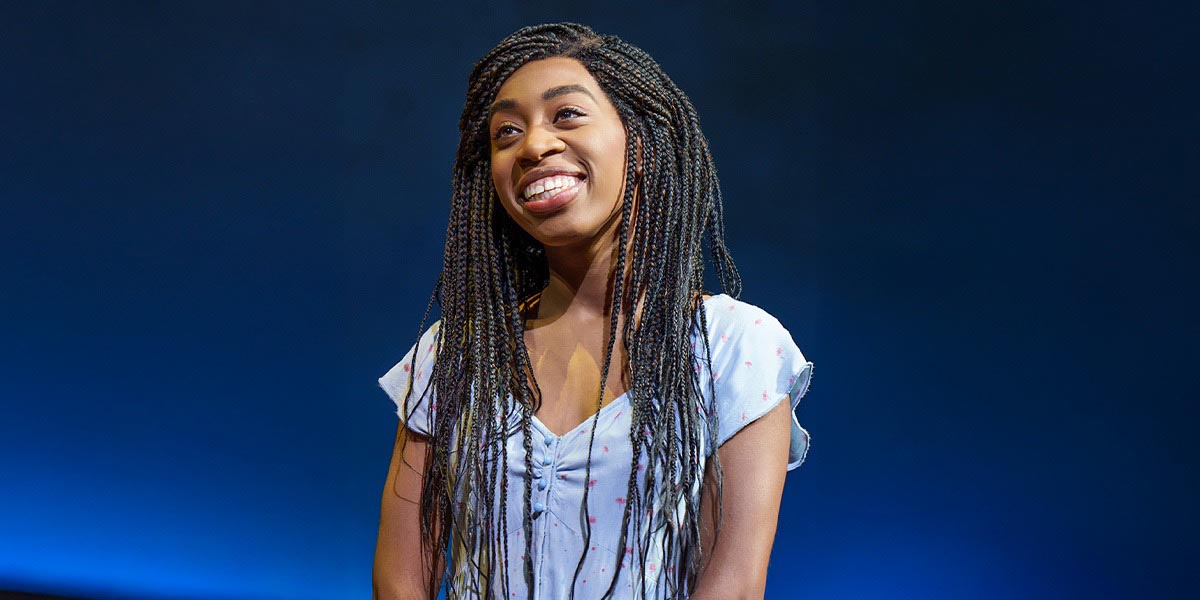 A smiling woman with long braided hair wears a light blue dress, against a dark blue background.