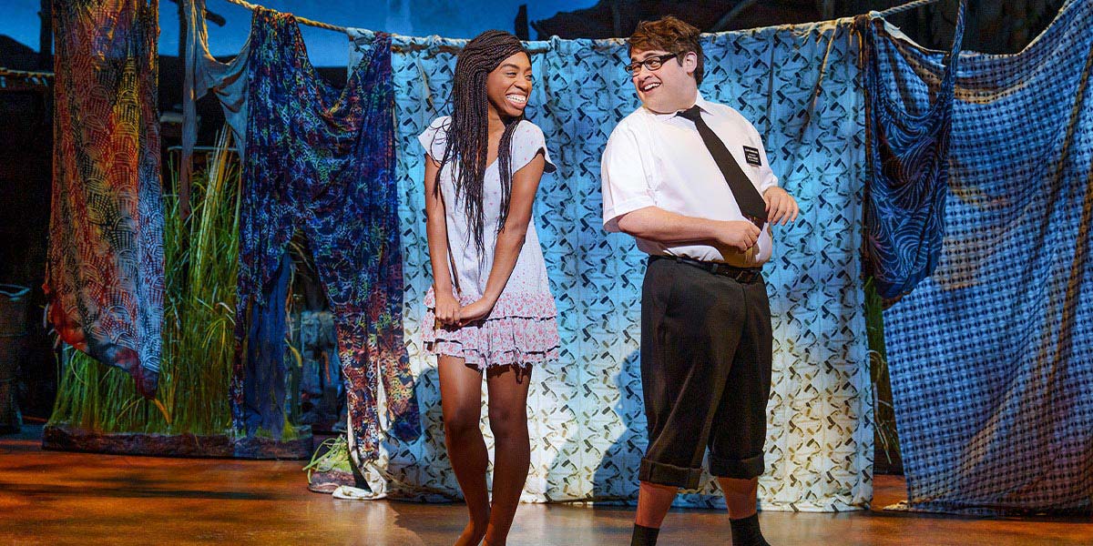 A scene from The Book of Mormon featuring performers in colourful costumes, singing on a stage with a vibrant backdrop.