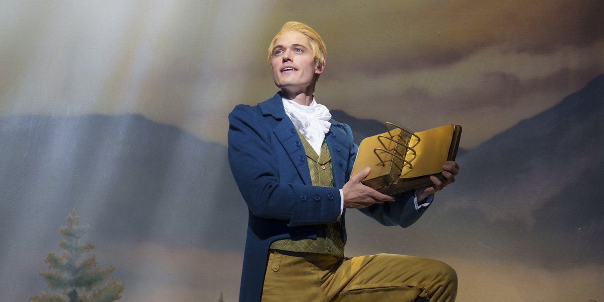 A man in a blue coat and yellow trousers holds a book, standing against a scenic backdrop.