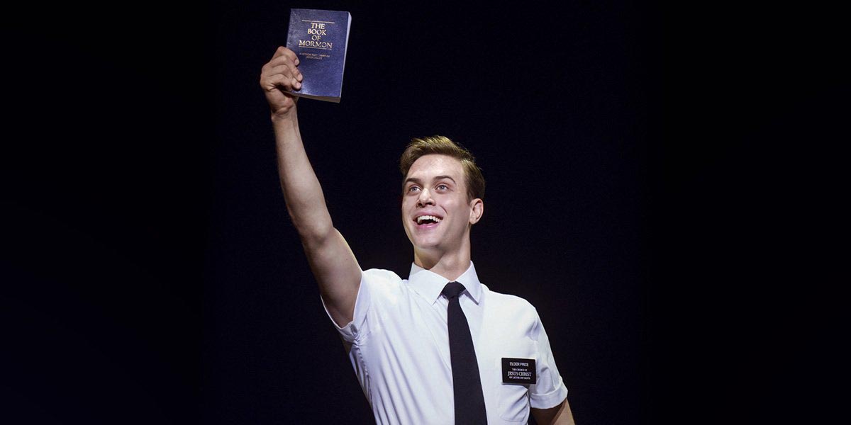 A smiling man in a white shirt and tie holds up a copy of "The Book of Mormon.