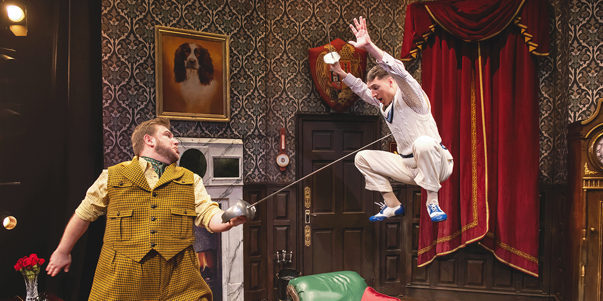 Two male actors in formal attire, one in a tuxedo and the other in a suit, perform on stage.