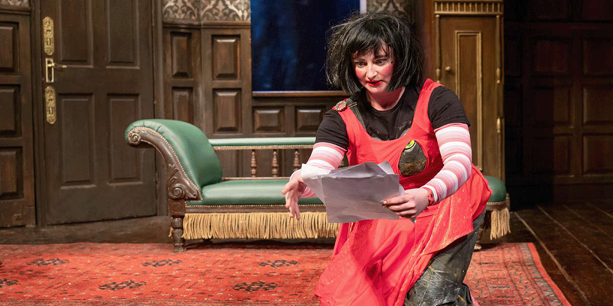 A woman with short hair, wearing a black dress, performs on stage with dramatic lighting and props.