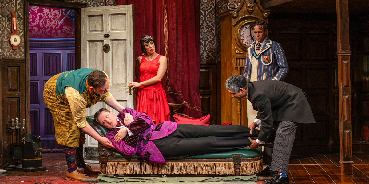 A group of actors in period costumes comically struggle with a large prop in a theatre setting.