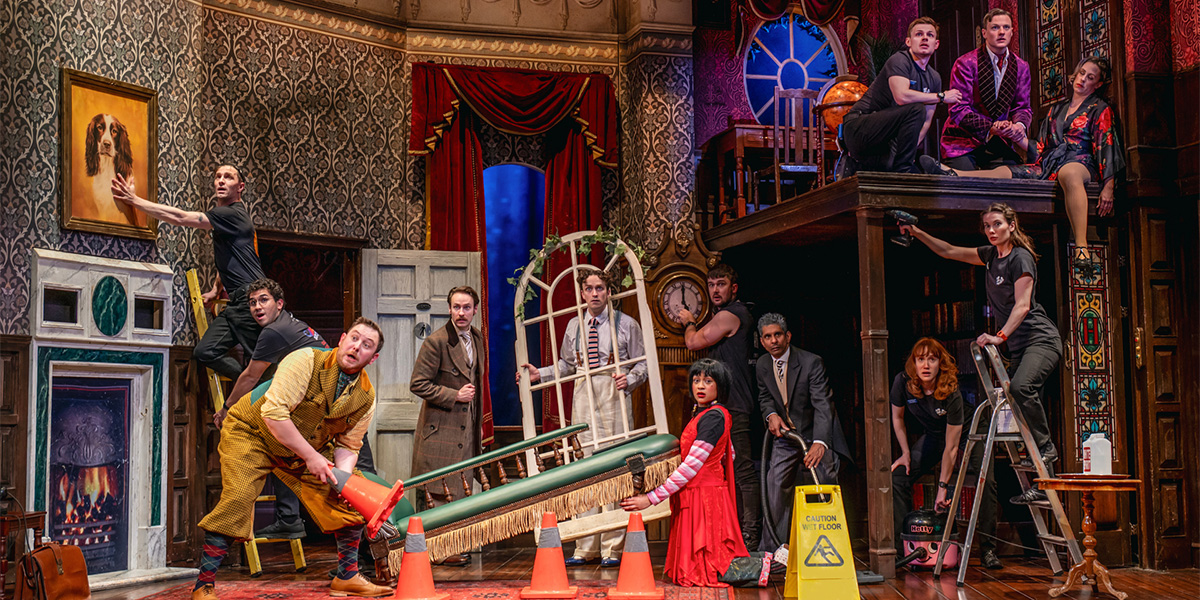 A group of actors from The Play That Goes Wrong perform a comedic scene on stage, with vibrant set design.