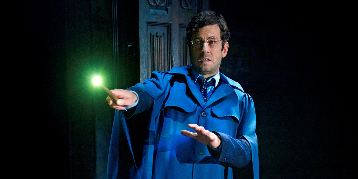 A man with glasses and a black robe stands on stage, surrounded by dramatic lighting and theatrical set pieces.