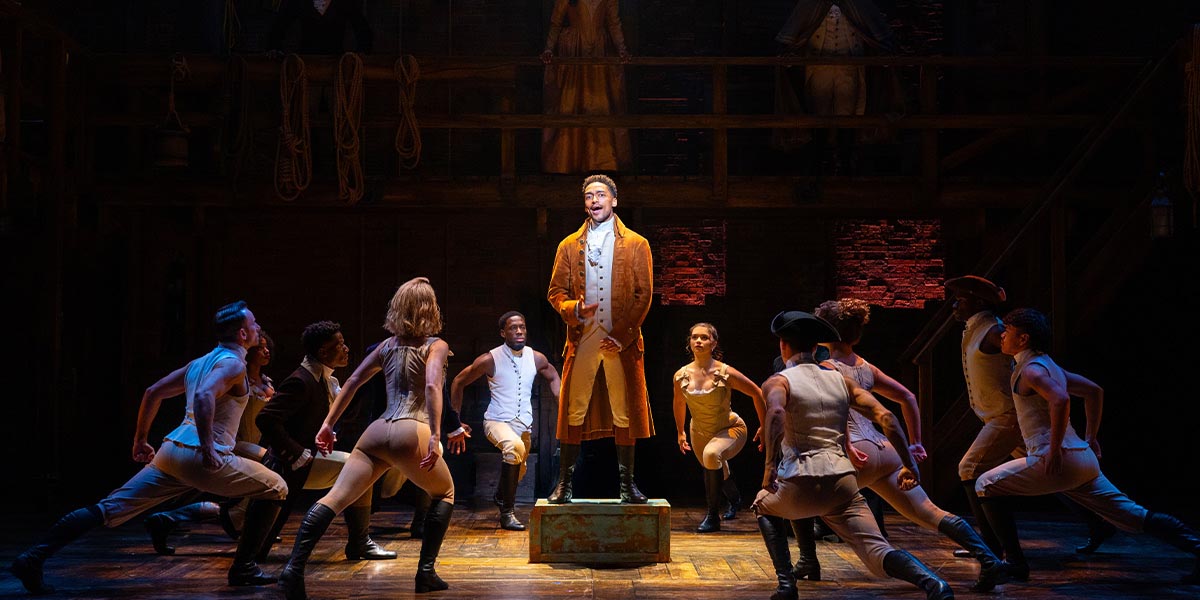 Alex Sawyer as Alexander Hamilton stands centre stage, surrounded by the ensemble, in period costumes.