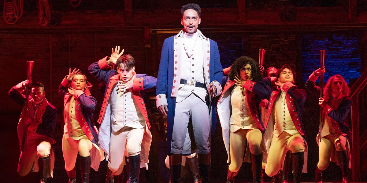 Alex Sawyer as Alexander Hamilton, wearing a historical costume, stands confidently against a dark background.