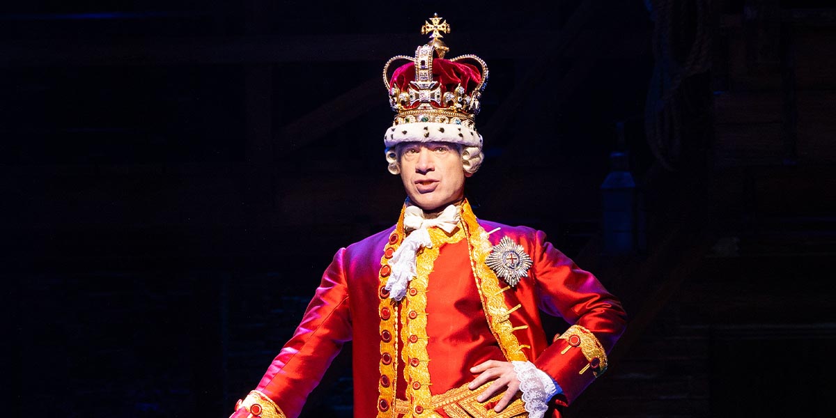 Daniel Boys as King George III, wearing a crown and red royal attire, striking a dramatic pose.