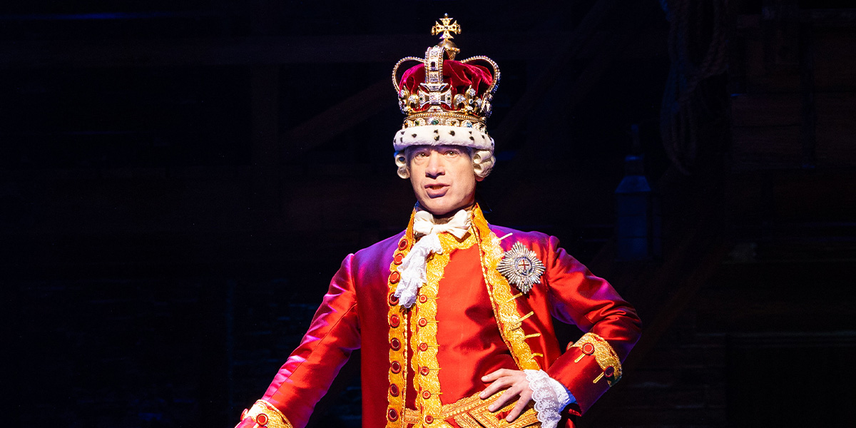 Daniel Boys as King George III, wearing a regal costume with a crown, smiling against a dark background.