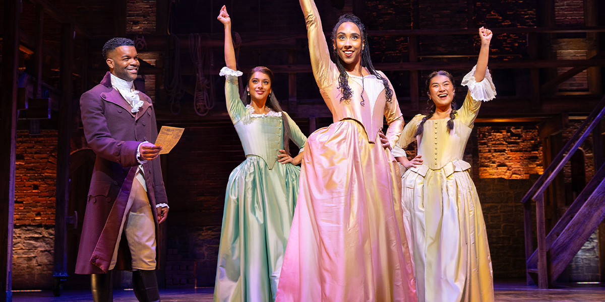 A group of actors in period costumes, portraying characters from Hamilton, poses together on stage.