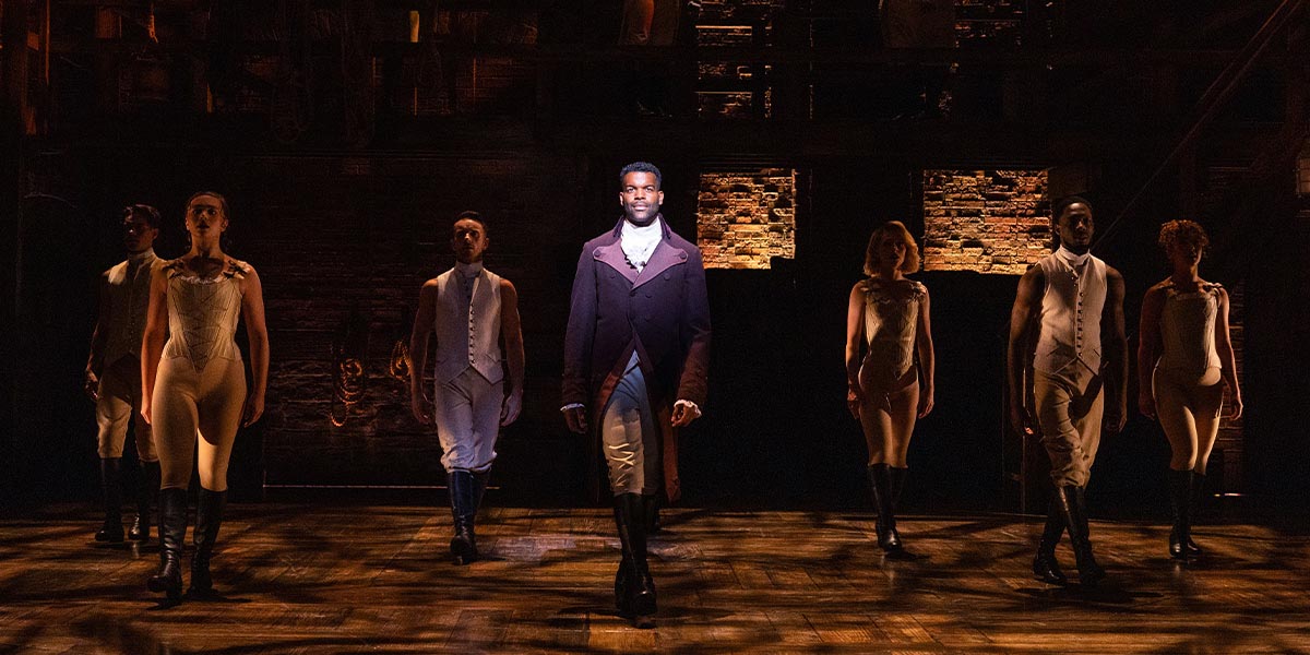 Jay Perry as Aaron Burr stands centre stage, surrounded by the ensemble in period costumes, dramatic lighting.