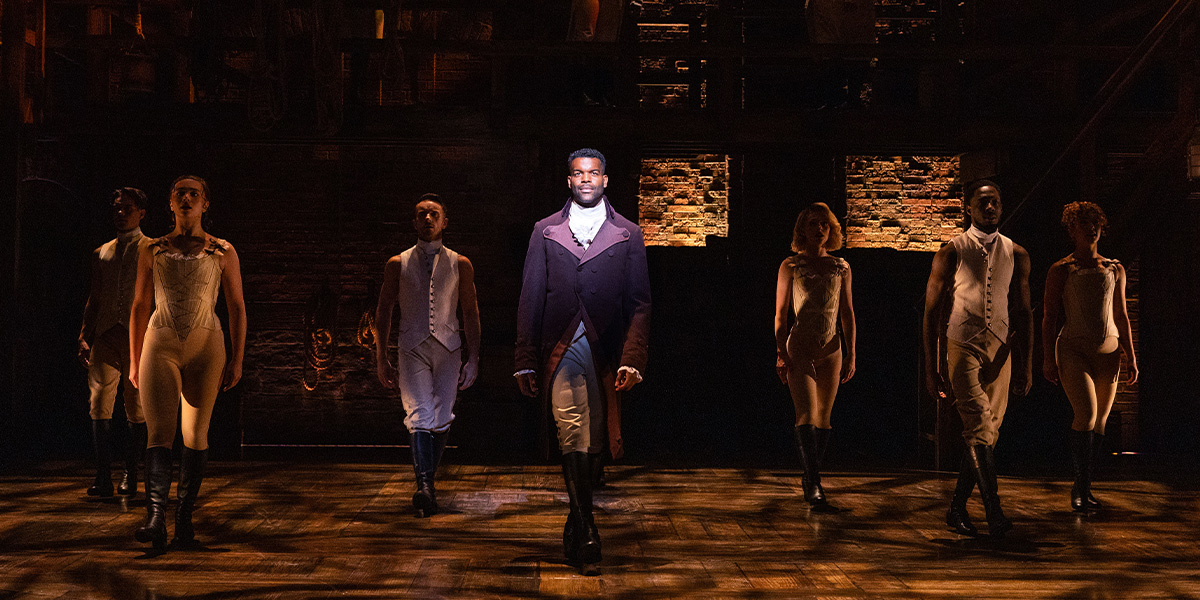 Jay Perry as Aaron Burr stands centre stage, surrounded by ensemble members in period costumes, dramatic lighting.