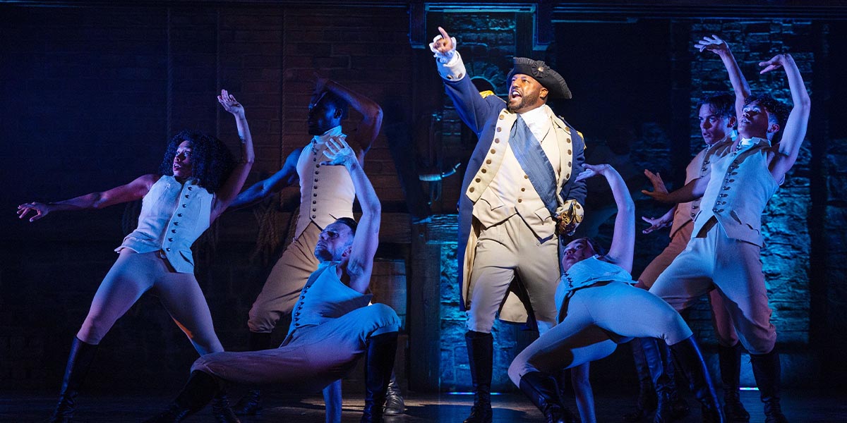 Jonathan Andrew Hume as George Washington stands centre stage, surrounded by cast members in period costumes.