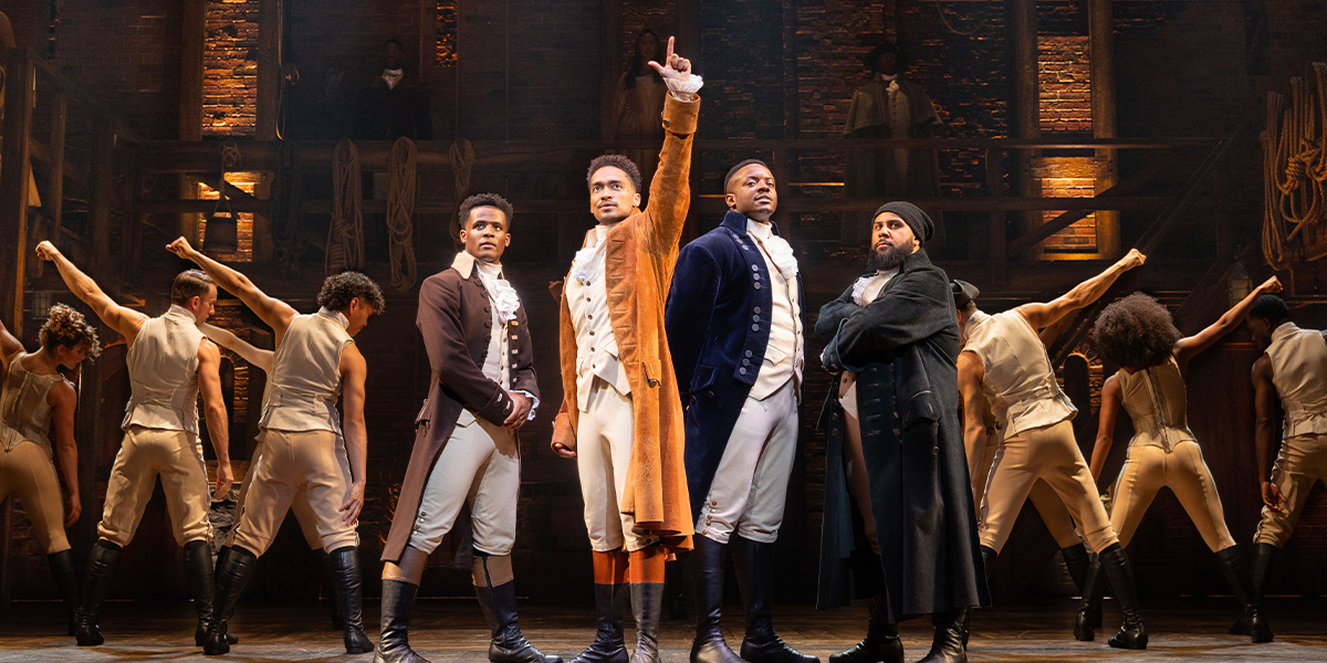 A group of actors in period costumes perform energetically on stage, with dramatic lighting and a dark backdrop.